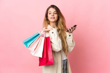 Alışveriş çantalı genç bir kadın pembe arka planda izole edilmiş. Elinde alışveriş çantaları ve cep telefonu var.