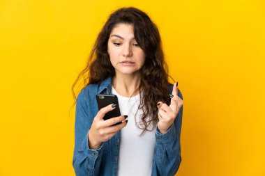 Sarı arka planda izole edilmiş genç Rus kız parmakları çapraz cep telefonu kullanıyor.