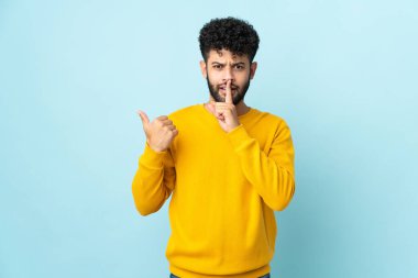 Faslı genç bir adam mavi arka planda izole edilmiş bir şekilde kenarı işaret ediyor ve sessiz hareketler yapıyor.