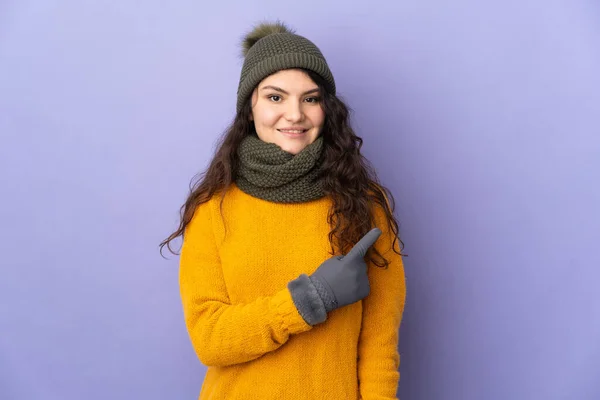 俄罗斯少女 头戴紫色背景的冬帽 指着侧面展示自己的产品 — 图库照片