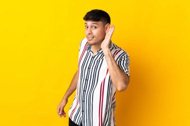 Genç Kolombiyalı adam sarı arka planda izole edilmiş. Kulağına elini sürerek bir şeyler dinliyor.