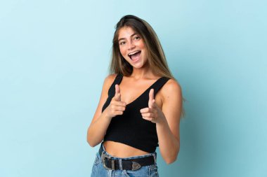 Mavi arka planda izole edilmiş genç beyaz kadın şaşırmış ve ön tarafı işaret ediyor.