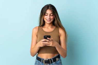 Mavi arka planda izole edilmiş genç beyaz kadın cep telefonuyla bir mesaj gönderiyor.