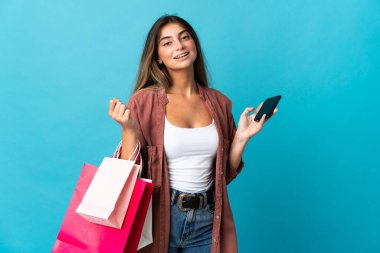 Kafkasyalı genç bir kadın mavi arka planda izole edilmiş. Elinde alışveriş poşetleri var ve bir arkadaşına cep telefonuyla bir mesaj yazıyor.