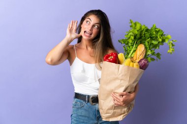 Market poşeti tutan genç bir kadın bir şey dinlerken elini kulağına koyar.