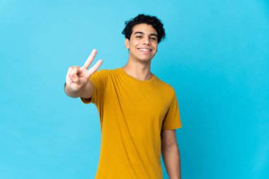 Genç Venezuelalı adam mavi arka planda izole edilmiş gülümsüyor ve zafer işareti gösteriyor.