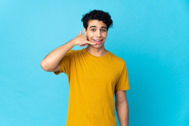 Genç Venezuelalı adam mavi arka planda izole edilmiş bir şekilde telefon jesti yapıyor. Bana geri dönüş işareti ver.