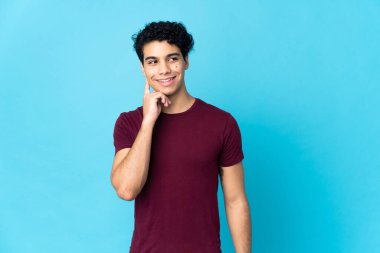 Genç Venezuelalı bir adam mavi arka planda izole edilmiş, yukarı bakarken bir fikir düşünüyor.