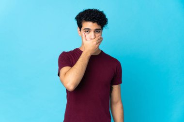 Genç Venezuelalı bir adam mavi arka planda izole edilmiş. Ağzını eliyle kapatıyor.