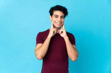 Genç Venezuelalı adam mavi arka planda izole edilmiş mutlu ve hoş bir ifadeyle gülümsüyor.