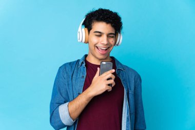 Genç Venezuelalı bir adam mavi arka planda izole edilmiş. Cep telefonuyla müzik dinliyor ve şarkı söylüyor.