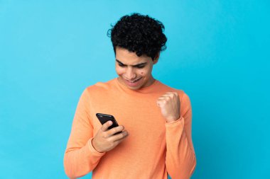 Venezuelalı adam mavi arka planda cep telefonu kullanarak ve zafer jesti yaparak izole edildi.