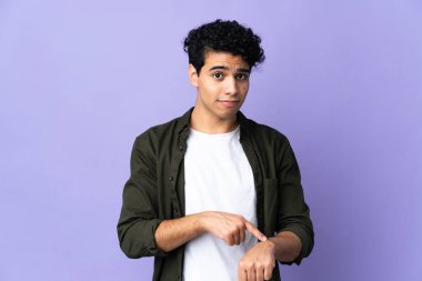 Genç Venezuelalı adam mor arka planda izole edilmiş, geç kalma jesti yapıyor.