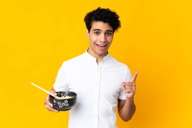 Genç Venezuelalı bir adam sarı arka planda izole edilmiş. Elinde çubuklarla bir kase erişte tutarken harika bir fikri işaret ediyor.