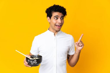 Genç Venezuelalı bir adam sarı arka planda izole edilmiş bir şekilde yemek çubuklarıyla bir kase erişteyi tutarken parmağını kaldırarak çözümü bulmaya çalışıyor.