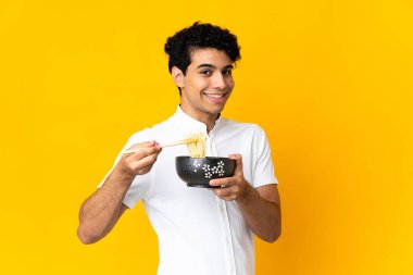 Genç Venezuelalı adam sarı arka planda izole edilmiş. Elinde çubuklu erişte kasesi var.