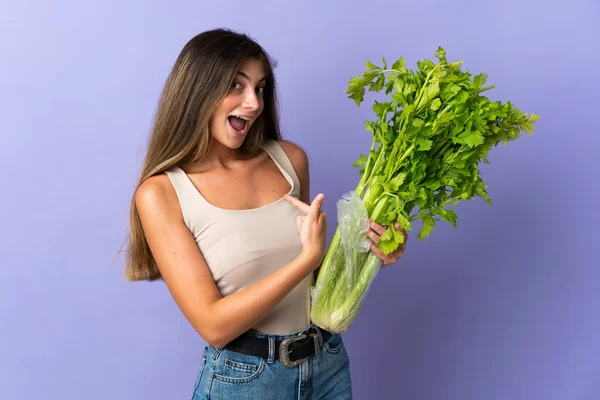 一个年轻的女人手里拿着一个紫色背景的芹菜 脸上带着惊讶的表情 — 图库照片