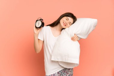 Genç Ukraynalı kız pembe arka planda pijamalarıyla izole edilmiş ve elinde mutlu bir ifadeyle saat ve yastık tutuyor.