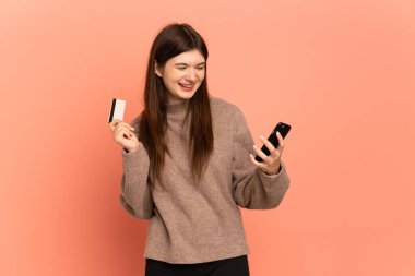 Pembe arka planda izole edilmiş genç Ukraynalı bir kız kredi kartıyla cep telefonuyla alışveriş yapıyor.