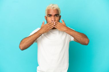 Genç Kolombiyalı yakışıklı adam mavi arka planda izole edilmiş ağzı elleriyle kapatıyor.