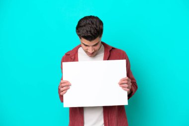 Genç, beyaz, yakışıklı bir adam mavi bir bakkal sahasında izole edilmiş, elinde boş bir levha var ve ona bakıyor.