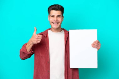 Genç, yakışıklı, beyaz bir adam mavi bir bakkalda izole edilmiş. Elinde başparmağıyla boş bir levha var.