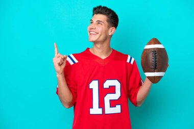 Genç beyaz adam rugby oynuyor. Mavi arka planda izole edilmiş. Harika bir fikre işaret ediyor.