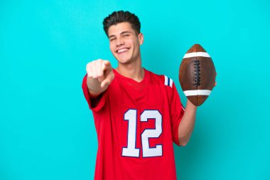 Genç beyaz adam rugby oynuyor mavi arka planda izole edilmiş, mutlu bir ifadeyle ön tarafı işaret ediyor.