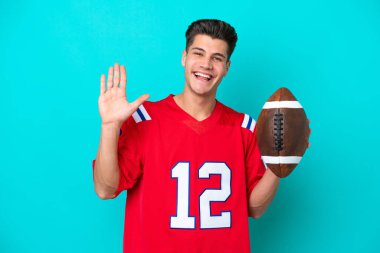 Genç beyaz adam rugby oynuyor. Mavi arka planda izole edilmiş. Mutlu bir ifadeyle selamlıyor.