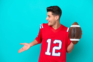 Rugby oynayan Kafkasyalı genç bir adam mavi arka planda yalıtılmış ve sürpriz bir ifadeyle yan tarafa bakıyor.