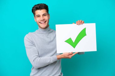Mavi bakkalda izole edilmiş genç beyaz bir adam elinde yazılı bir pankart tutuyor yeşil işaret simgesi ve mutlu bir ifade.