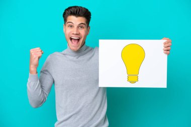 Genç, yakışıklı, beyaz bir adam mavi bir bakkalda izole edilmiş. Elinde ampul simgesi olan bir pankart var ve zaferi kutluyor.