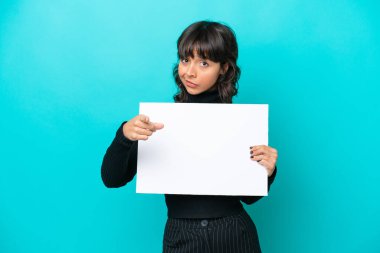 Genç Latin bir kadın mavi arka planda izole edilmiş elinde boş bir levha var ve ön tarafı işaret ediyor.