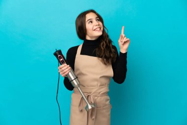 Mavi arka planda izole edilmiş el blenderi kullanan küçük kız harika bir fikre işaret ediyor.