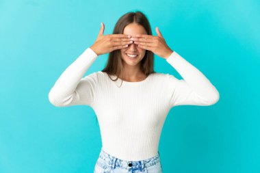 İzole edilmiş mavi arka planda genç bir kadın. Gözleri elleriyle kaplıyor ve gülümsüyor.