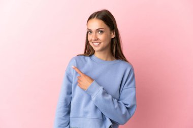 Soyutlanmış pembe arka planı olan genç bir kadın bir ürünü sunmak için kenarı işaret ediyor.