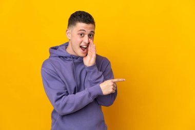 Sarı arka planda izole edilmiş genç beyaz bir adam bir ürün sunmak ve bir şeyler fısıldamak için kenarı işaret ediyor.