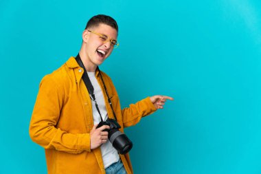 Kafkasyalı genç fotoğrafçı, mavi arka planda izole edilmiş bir şekilde parmak ucunda bir ürün sunuyor.