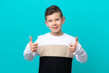 Küçük çocuk mavi arka planda izole edilmiş. Baş parmakları havada ve gülümsüyor.