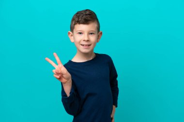 Küçük çocuk mavi arka planda izole edilmiş gülümsüyor ve zafer işareti gösteriyor.