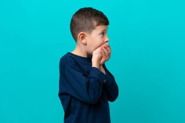 Küçük çocuk mavi arka planda izole edilmiş ağzını kapatıyor ve yan tarafa bakıyor.