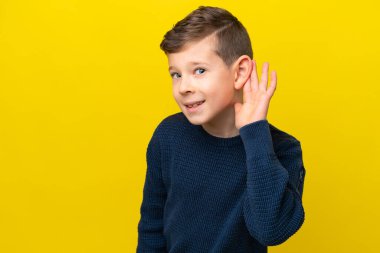 Küçük beyaz çocuk sarı arka planda izole edilmiş bir şekilde elini kulağına koyarak bir şeyler dinliyor.