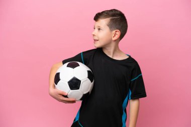 Küçük beyaz futbolcu çocuk pembe arka planda izole edilmiş, yan tarafa bakıyor ve gülümsüyor.