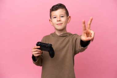 Pembe arka planda izole edilmiş bir bilgisayar oyunu kontrolörüyle oynayan beyaz çocuk gülümsüyor ve zafer işareti gösteriyor.