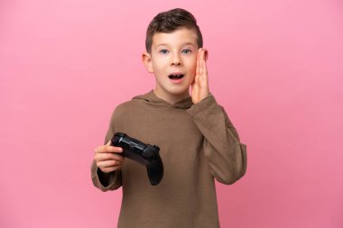 Küçük beyaz çocuk pembe arka planda izole edilmiş bir bilgisayar oyunu kontrolörüyle oynuyor. Şaşırmış ve şok olmuş yüz ifadesi var.