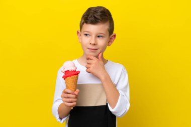 Sarı arka planda izole edilmiş kornet dondurması olan küçük beyaz çocuk yana bakıyor ve gülümsüyor.