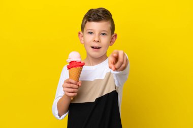 Kornet dondurması olan küçük beyaz çocuk sarı arka planda izole edilmiş şaşırmış ve ön tarafı işaret ediyor.