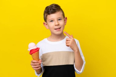 Kornet dondurması olan küçük beyaz bir çocuk sarı arka planda izole edilmiş bir şekilde baş parmak hareketi yapıyor.