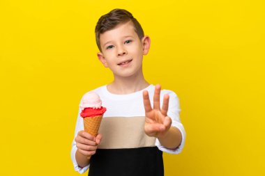 Kornet dondurmalı küçük beyaz çocuk sarı arka planda izole edilmiş mutlu ve parmaklarıyla üçe kadar sayıyor.