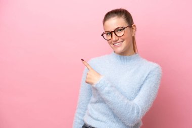 Pembe arka planda izole edilmiş genç beyaz kadın bir ürünü sunmak için kenarı işaret ediyor.